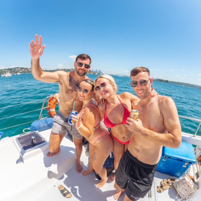 Four adults in swimsuits smile and pose together on a boat under a sunny sky, each holding a drink. The ocean and a distant city skyline are visible in the background.