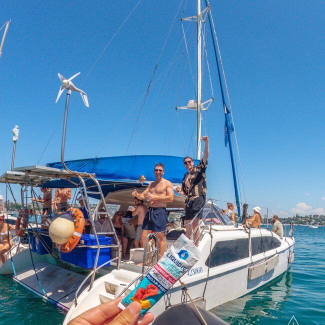 A group of people are enjoying a sunny day on a white sailboat anchored on blue water. In the foreground, a person holds a flavored electrolyte gel pack. The sky is clear and the mood is festive.
