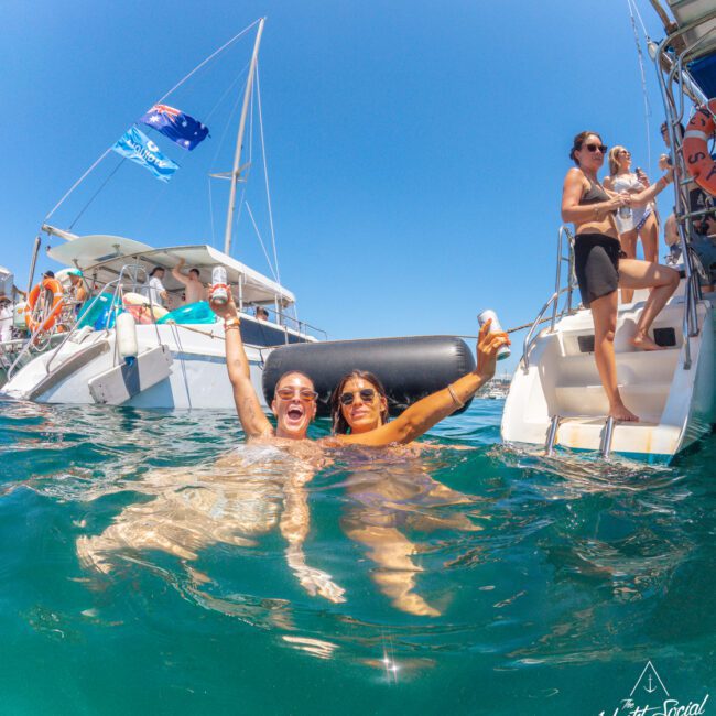 Two smiling women float in the ocean holding drinks near a yacht, surrounded by other people on boats under a clear blue sky, with an Australian flag visible.