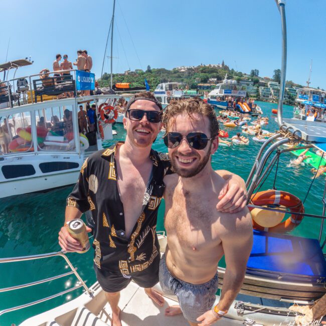 Two men in swimwear and sunglasses smile on a boat, surrounded by other boats and people swimming in clear blue water under a sunny sky, creating a lively summer party atmosphere.