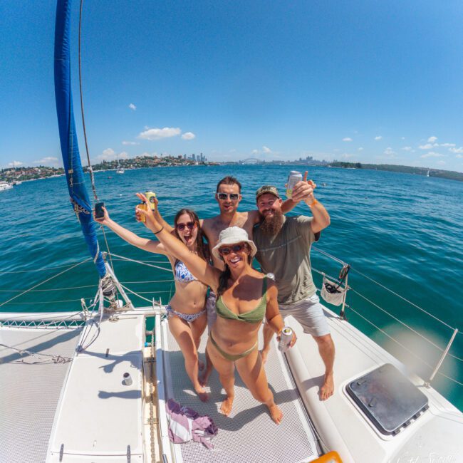 Four people in swimsuits smiling and raising drinks while posing on a sailboat deck, with blue water and a city skyline visible in the background under a clear, sunny sky.