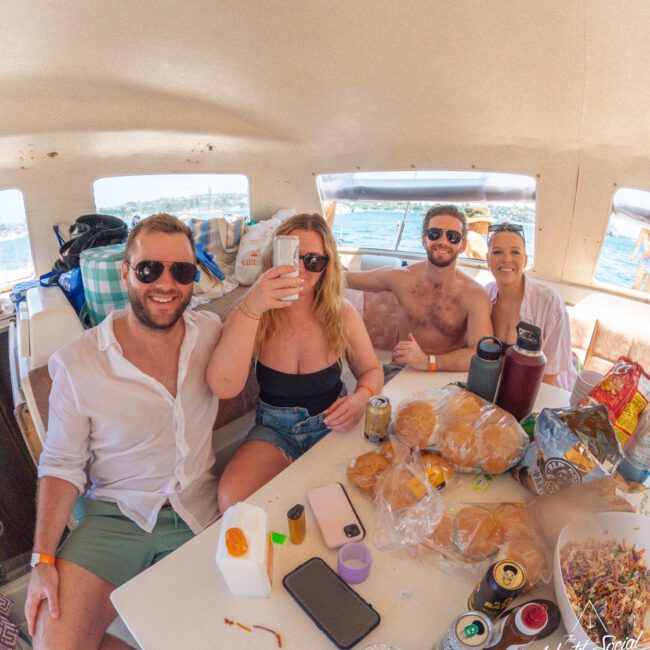 Four people in swimwear and light clothing sit around a table with food and drinks on a boat, smiling and raising drinks, with water and blue sky visible through the windows.