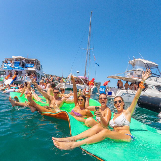 A group of people in swimsuits relax and wave on large green floats in the water, with boats and a clear blue sky in the background. Everyone looks happy and festive, enjoying a sunny day at a boat party.