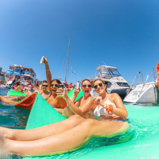A group of women in swimsuits relax on a floating mat in the water, smiling and holding drinks, with boats and other people partying in the background under a clear blue sky.