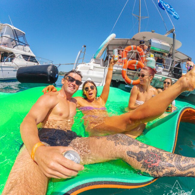 Three people relax on a green float in the water, smiling and raising drinks. Boats and a clear blue sky are in the background, suggesting a fun, sunny day on the water.