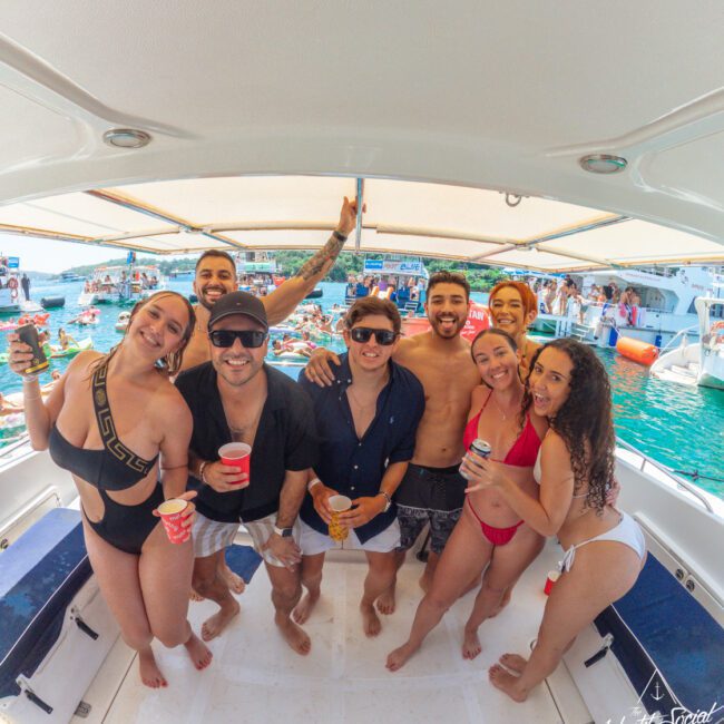 A group of seven young adults smiling and holding drinks poses on a boat deck, with people, boats, and blue water visible in the sunny background. It looks like a lively party scene.
