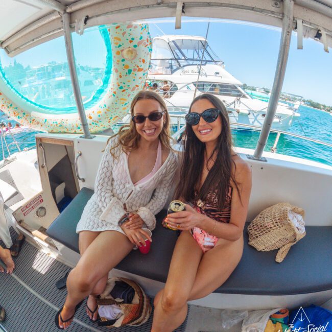 Two women wearing swimsuits and sunglasses sit smiling on a boat bench, holding drinks. A sunny day, with yachts and water in the background, and a large inflatable float hanging nearby.