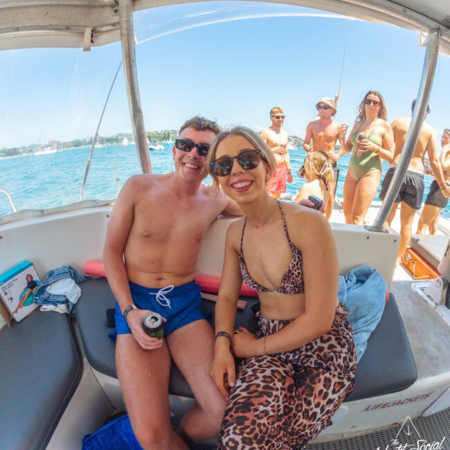 Two people in swimwear smile and sit close together on a boat, with a group of people standing and socializing in swimsuits on the deck behind them. It's a sunny day on the water.