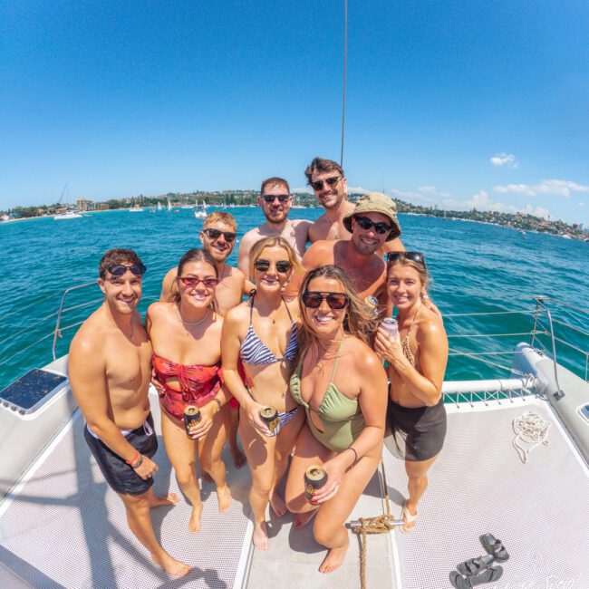 A group of ten smiling people in swimsuits stand together on a boat deck, holding drinks, with bright blue water and a sunny sky in the background.
