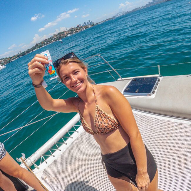 A smiling woman in a leopard-print bikini top and black shorts stands on a boat, holding up a card. The ocean and city skyline are visible in the background under a sunny blue sky.