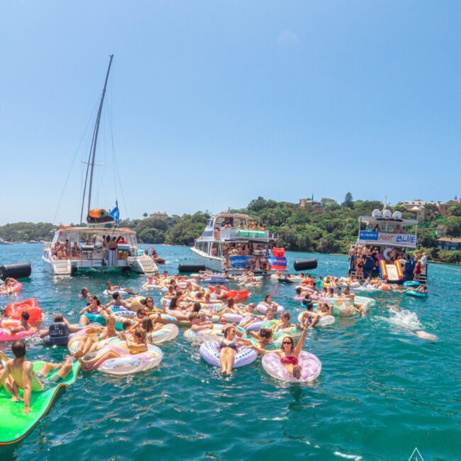 A group of people relax on inflatable tubes in the water near anchored boats on a sunny day. The participants are enjoying a lively outdoor party, surrounded by green hills and clear blue sky.