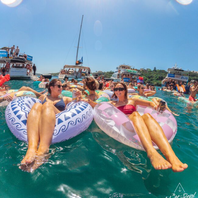 Two women relaxing on inflatable pool floats, smiling and holding drinks, surrounded by other people in the water with boats and a clear blue sky in the background. The scene is lively and summery.