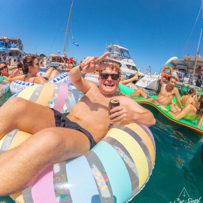 A man in swim trunks smiles and flashes a peace sign while lounging on a colorful inflatable ring in the water; boats and other people on floaties are visible under a clear blue sky. The Yacht Social Club logo appears in the corner.