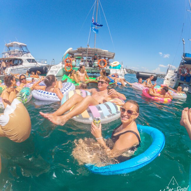 A group of people relaxing on colorful pool floats in the ocean near yachts, enjoying drinks and sunny weather at a social boat party. Blue sky and boats are visible in the background.