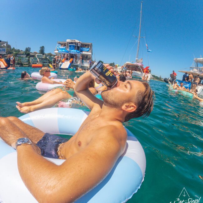 A man lounges on a pool float in clear blue water, drinking from a can. Other people relax on floats nearby, with boats and more people in the background under a sunny, blue sky.