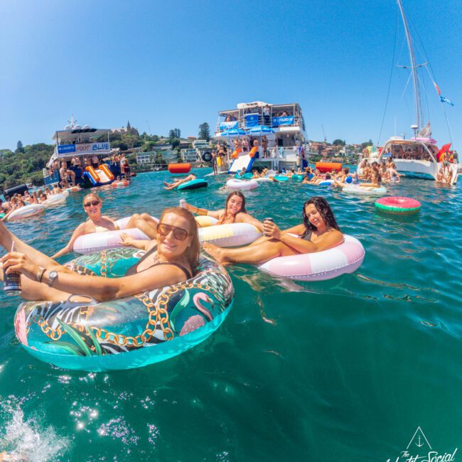 A group of people relax and smile on colorful inflatable tubes in clear blue water, surrounded by boats and others enjoying a sunny day at a lively social gathering on the water.