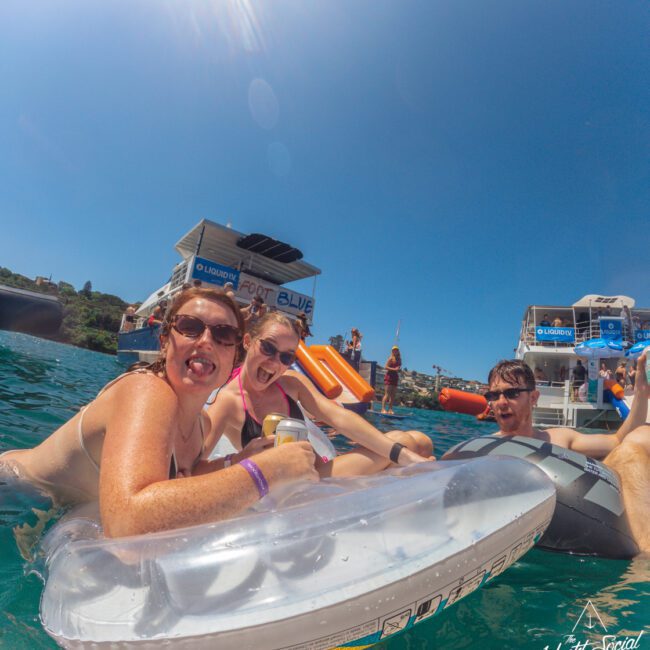 Three people are smiling and making faces while relaxing on inflatable floats in clear blue water, with a large boat and water slide in the background under a sunny sky.