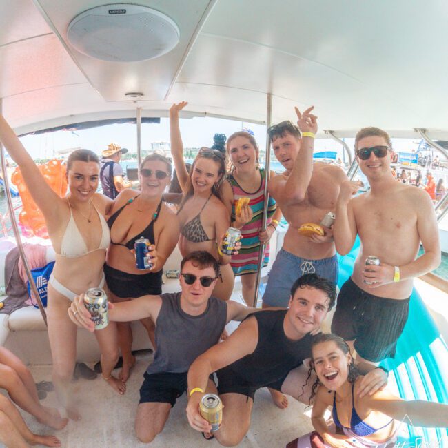 A group of young adults in swimwear smile and pose with drinks on a boat, celebrating under a canopy. Some are raising their arms and others are sitting or kneeling, enjoying a sunny day on the water.