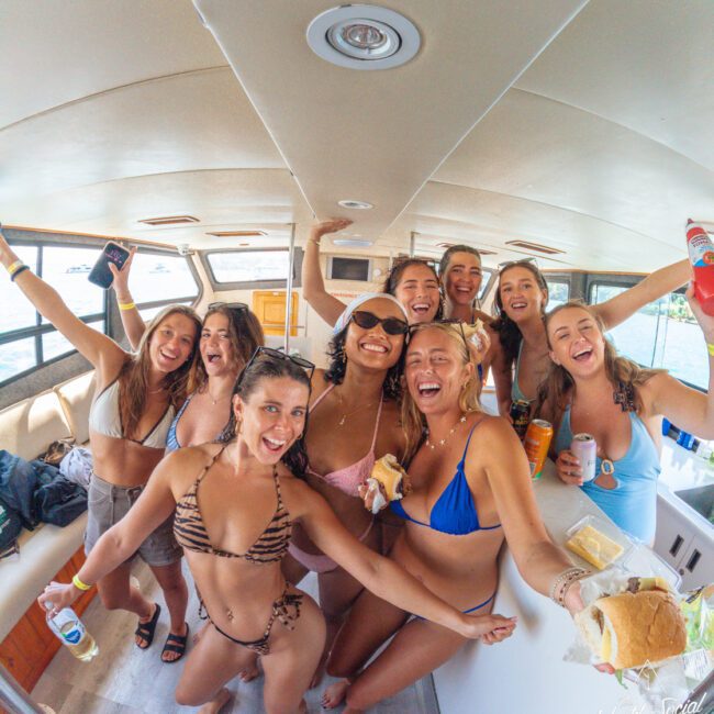 A group of women in swimsuits laugh and pose together on a boat, holding drinks and sandwiches, enjoying a fun day on the water. The mood is joyful and festive.