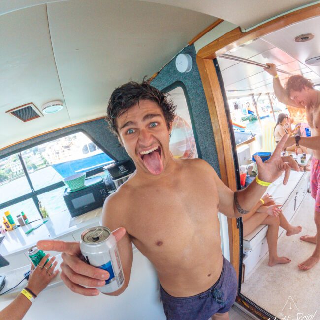 A shirtless young man with wet hair stands on a boat, smiling and sticking out his tongue, holding a can and gesturing with his hand. Other people are in the background, enjoying the party atmosphere.