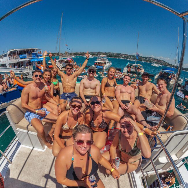 A group of young adults in swimwear smile and pose together on a boat under sunny skies, surrounded by other boats on a lively, blue body of water. Some hold drinks, enjoying a festive, summer atmosphere.