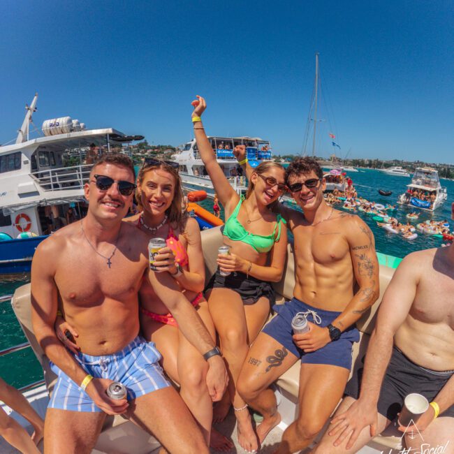 Four young adults in swimwear sit on a boat, smiling and holding drinks, with one woman raising her arm in excitement. Behind them, other boats and people are enjoying a sunny day on the water.