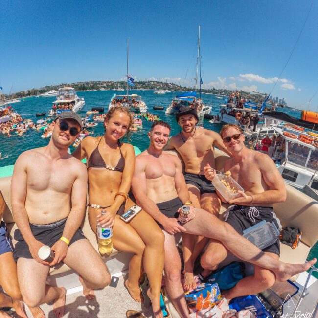 A group of five friends in swimwear relax and smile on a boat, with crowded party boats and people swimming in the blue water under a sunny sky in the background.