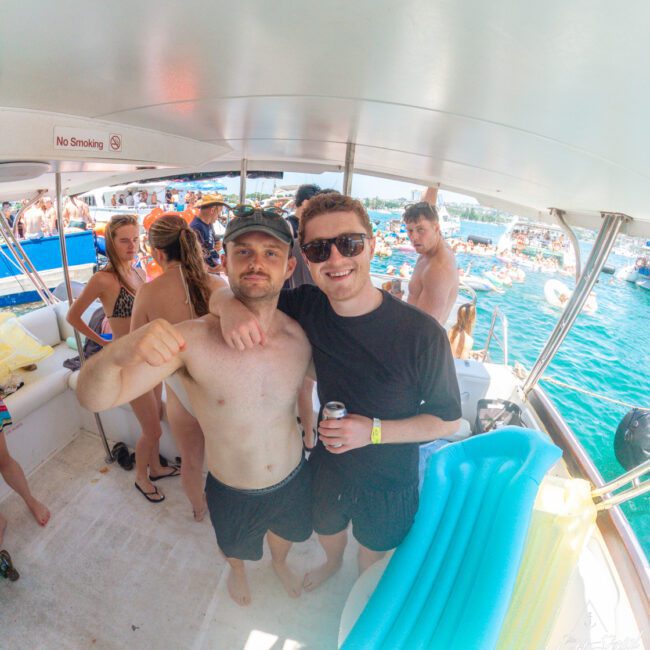 Two men in swim trunks pose smiling on a boat, one with his arm around the other. Other people in swimwear enjoy the boat and water in the background, with inflatables and a blue sky visible.