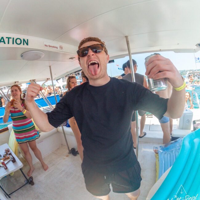 A man in sunglasses and black clothing stands on a boat, holding a drink and sticking out his tongue playfully. Other people are around him, enjoying the festive atmosphere on a sunny day.