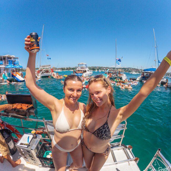 Two women in bikinis stand on a boat smiling with arms raised, one holding a drink. Behind them, people swim and relax on other boats in a sunny, crowded marina. A man grills food on the left. The atmosphere is lively and festive.