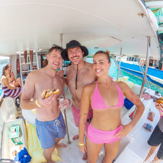 Three people in swimsuits smile and pose on a boat; one holds food, another wears a black hat. Other people and a blue boat are visible in the background, with sunny weather and water nearby.