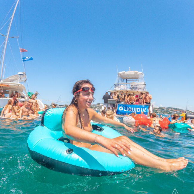 A woman in sunglasses floats on a blue inflatable tube in the ocean, smiling, with yachts and a group of people partying in the background under a clear blue sky.