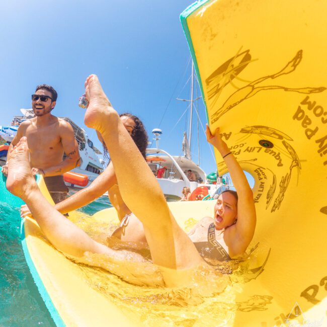 A woman joyfully falls off a yellow floating pad into the water, with laughter and splashes around her. Two people in swimsuits stand nearby, smiling, and a yacht is visible in the sunny background.