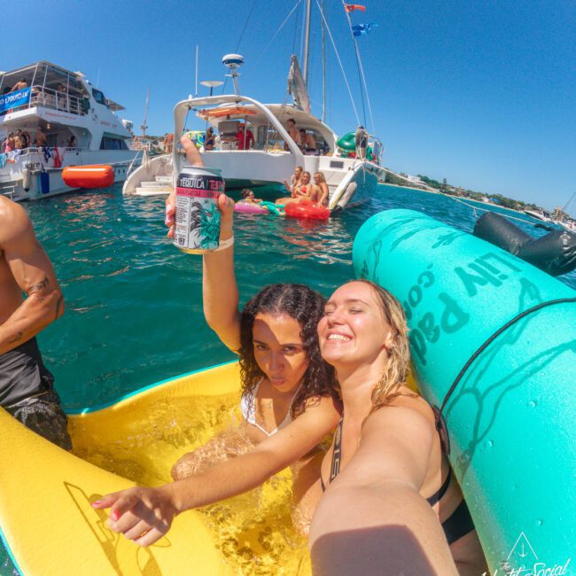 Two women on a yellow float smile and pose for a selfie in the water near yachts. One woman holds up a can, and people relax on boats and floats in the background under a sunny blue sky.