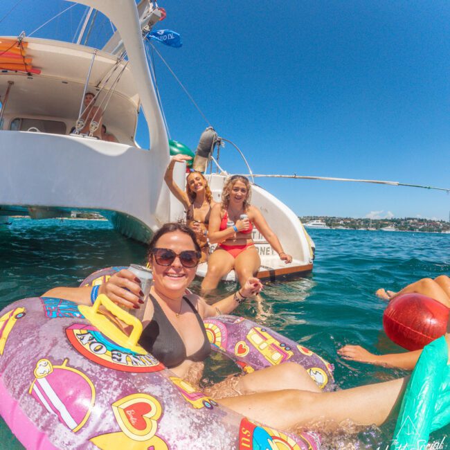 Four women enjoy a sunny day on a boat; one relaxes on a colorful float in the water, holding a drink, while three others sit on the boat’s edge, smiling, with clear turquoise water and a coastal view in the background.