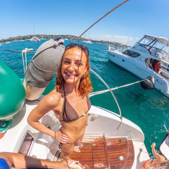 A smiling woman in a brown bikini stands on a boat deck under bright sun, with blue-green water and another boat in the background. The scene is lively and cheerful, suggesting a fun day on the water.