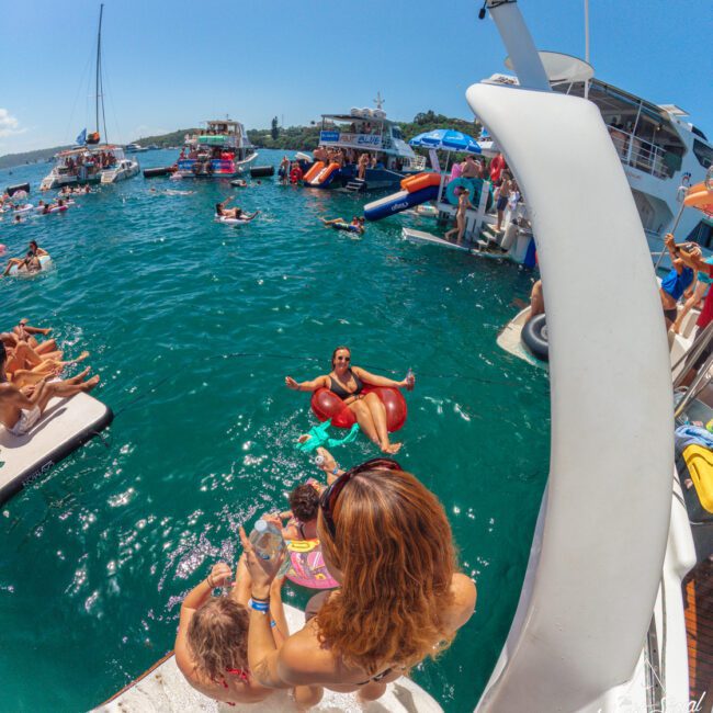 People enjoy a lively boat party, relaxing on floats and swimming in clear blue water. A woman helps a child at the top of a slide, while another person splashes into the sea below. Boats are anchored nearby under a sunny sky.