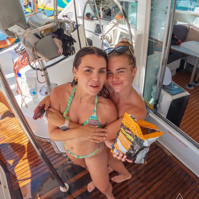 Two women in swimsuits stand on a yacht, smiling at the camera. One holds a bag of chips. The scene is bright and sunny, with nautical equipment and a wooden deck visible. The logo “Yacht Social Club” appears in the corner.