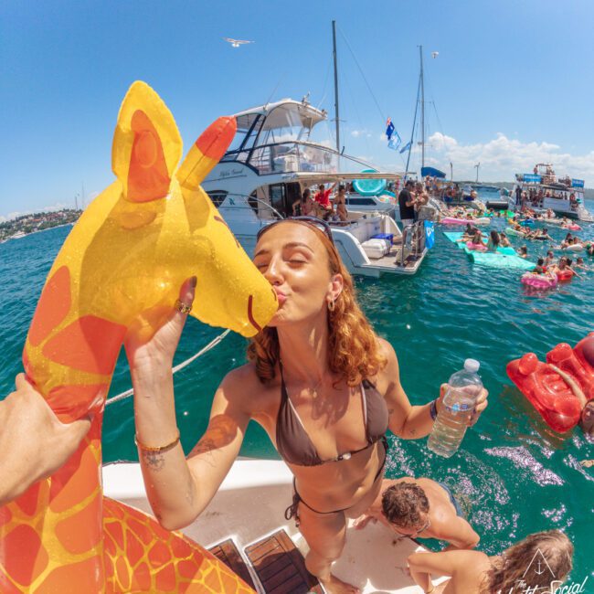 A woman in a bikini kisses an inflatable giraffe on a boat surrounded by people swimming and relaxing on floats in the water, with other boats and a sunny sky in the background.