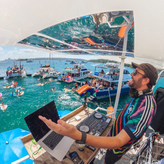 A DJ in colorful clothes performs on a boat deck overlooking a lively party with people swimming and relaxing on boats in a sunny, blue waterfront setting. Reflections of the scene are visible above him.