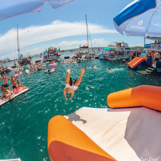 A person does a backflip off an orange and white inflatable slide into the ocean, surrounded by boats and people enjoying a sunny day on the water.