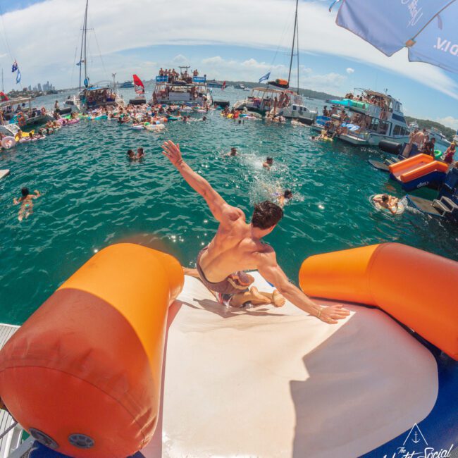 A man slides down an inflatable slide from a boat into the ocean, surrounded by people swimming, lounging on floats, and several boats anchored nearby under a sunny sky.