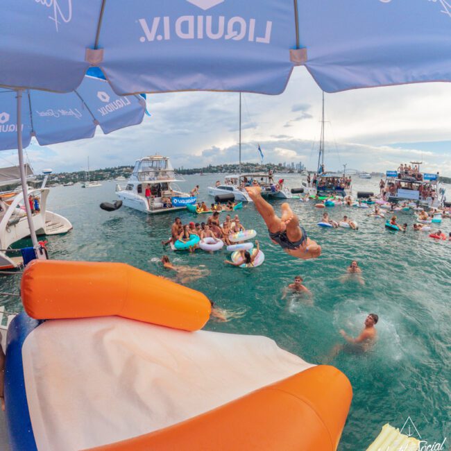 People swim and float in a busy bay filled with boats. A person dives into the water from an inflatable slide under a large branded umbrella. The sky is cloudy, and there are yachts and city buildings in the distance.
