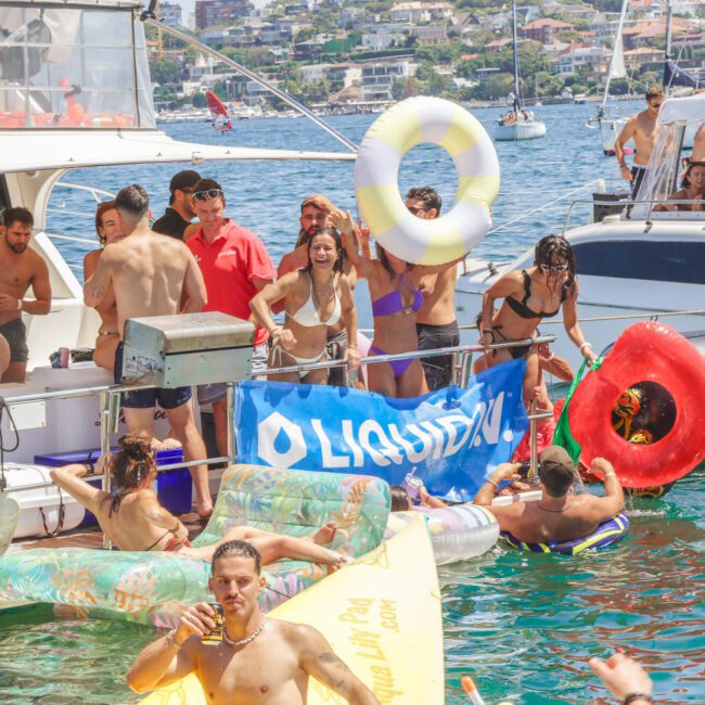 A group of people in swimwear enjoy a lively boat party on a sunny day, holding inflatable rings and floating on rafts in the water near two yachts, with a blue "LIQUID" banner displayed.