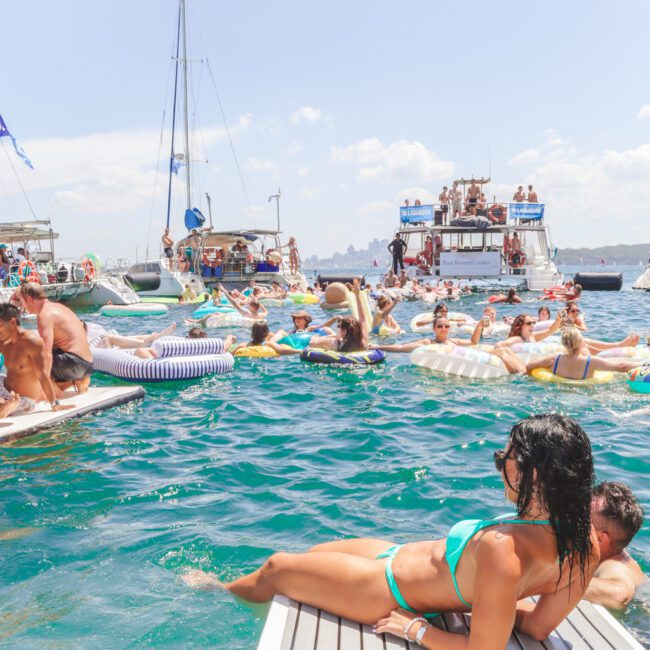 People relax on inflatables and boats in a crowded, sunny party scene on the water. A woman in a turquoise bikini lounges on a dock in the foreground, with yachts and swimmers in the background.