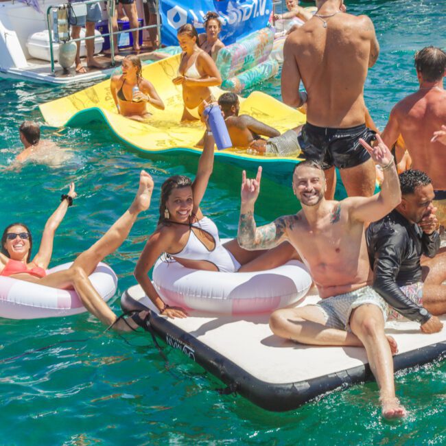 A group of people in swimsuits enjoy a sunny day on the water, lounging on inflatable rafts and floating mats, laughing, smiling, and posing for the camera in the bright blue sea.