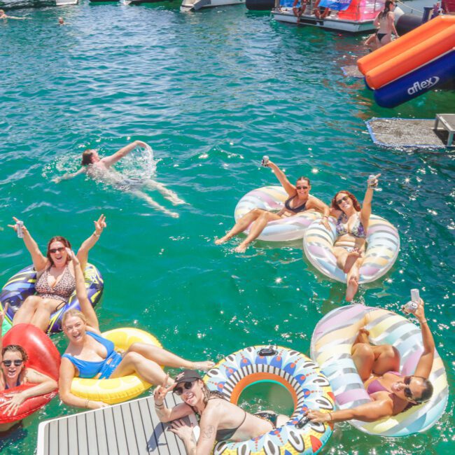 A group of people in swimsuits relax and smile on colorful inflatable floats in a lake, with more people and boats in the background. Some are waving and posing for the camera in bright sunlight.