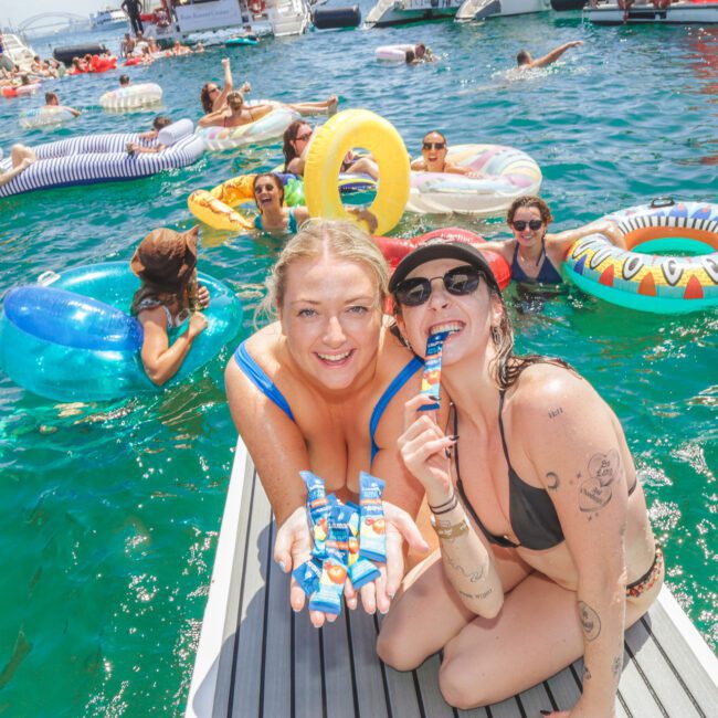 Two smiling women on a dock hold drink pouches, surrounded by people in colorful pool floats enjoying a sunny day on the water with boats in the background.