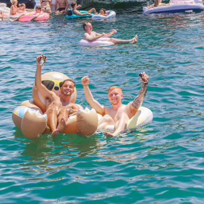 Two men on an inflatable emoji float raise drinks and smile at a crowded pool party, surrounded by others relaxing on colorful floaties in the bright blue water.