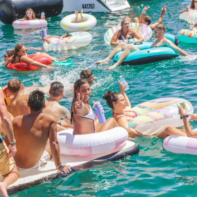 A group of people enjoying a sunny day on the water, relaxing on colorful inflatable floaties and boats. Some are swimming, others are lounging, laughing, and holding drinks, creating a lively and festive atmosphere.